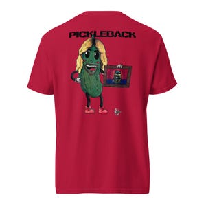 May include: A red t-shirt with the word "PICKLEBACK" printed in black. The shirt features a cartoon pickle character with blonde hair, holding a framed picture of a pickle jar. The pickle is wearing red shoes.
