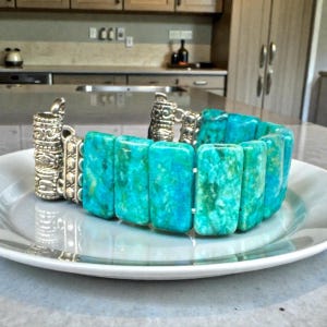 May include: A turquoise and silver bracelet with rectangular turquoise stones. The bracelet is displayed on a white plate. The silver clasps have an ornate design. The turquoise stones have a marbled appearance.