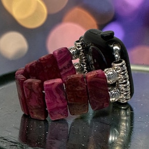 May include: A watch band featuring rectangular, marbled magenta and deep red stone beads connected by ornate, silver-toned metal spacers. The band attaches to a black smartwatch housing, showcasing a textured, multi-bead design with metallic accents.