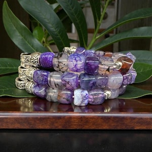May include: A stack of three bracelets with purple, gray, and brown beads. The beads are square-shaped and have a marbled appearance. The bracelets have silver-colored metal accents and are displayed on a wooden surface with green leaves in the background.