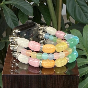 May include: A multi-strand bracelet with a variety of colorful beads, including white, pink, yellow, orange, and green, is paired with a black smartwatch. The bracelet features silver-toned metal accents and is displayed on a dark wooden surface.