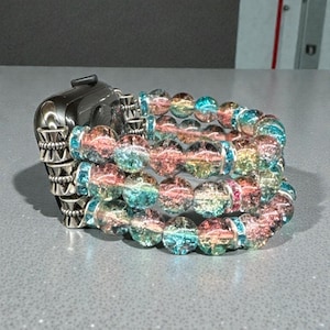 May include: A stack of three bracelets with iridescent, colorful beads. The beads transition through shades of pink, yellow, and blue. The bracelets are next to a silver watch.