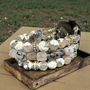 May include: Three stacked bracelets with white, gray, and black marbled beads and silver-colored metal accents. The bracelets are displayed on a small wooden tray. A black bottle is behind the bracelets.