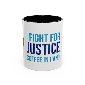 May include: White ceramic coffee mug with a black interior and the text "I FIGHT FOR JUSTICE COFFEE IN HAND" in blue and teal. A brown fist graphic is on the left side of the mug.
