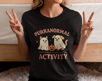 May include: Black t-shirt with the text "PURRANORMAL ACTIVITY" in peach-coloured letters. The graphic features a cat and a dog dressed as ghosts, with a pumpkin in the middle. The shirt is made of a soft material.