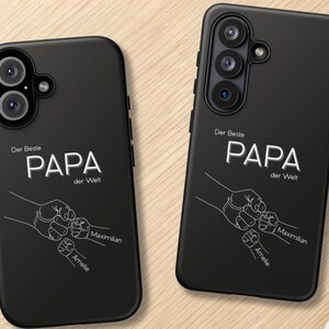 Könnte beinhalten: Zwei schwarze Handyhüllen mit dem Text "Der Beste PAPA der Welt" und einer Grafik von Händen, die sich halten. Die Namen "Maximilian" und "Amelie" stehen darunter. Die Hüllen liegen auf einer hellbraunen Holzoberfläche.