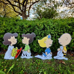 May include: Four Peanuts character cutouts standing in a row. The first figure has dark hair and a light blue gown, the second has dark hair and a green shirt, the third has blonde hair and a blue dress, and the fourth is a bald figure in a yellow shirt.
