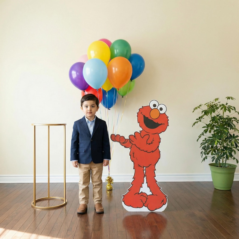 May include: A young person in a navy blue blazer and khaki pants stands next to a large Elmo cutout and a bunch of colorful balloons. A gold side table and a potted plant are also in the room. The balloons are in various colors, including blue, yellow, and green.