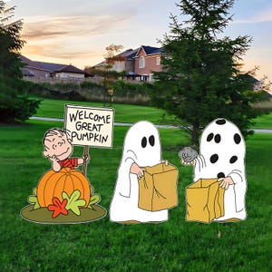 May include: A set of outdoor yard decorations featuring Linus from Peanuts sitting behind a large orange pumpkin with a sign that reads WELCOME GREAT PUMPKIN, alongside two figures dressed as ghosts holding brown paper trick-or-treat bags.