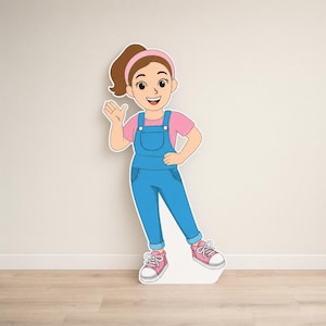May include: A full-length cutout of a cartoon girl wearing a pink t-shirt, blue dungarees, pink trainers, and a pink headband. The girl is waving with her right hand and smiling. The cutout is standing on a wooden floor against a beige wall.