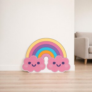 May include: A decorative rainbow and cloud cut-out. The rainbow has bands of yellow, pink, purple, blue, and orange. Two pink clouds with smiling faces sit below. It's positioned against a white wall and wooden floor.