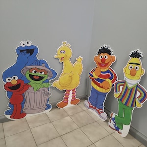 May include: Cardboard cutouts of Sesame Street characters, including Cookie Monster, Elmo, Oscar the Grouch, Big Bird, Bert, and Ernie. The characters are brightly colored and stand against a gray wall.