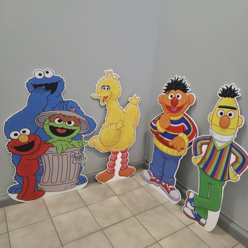 May include: Cardboard cutouts of Sesame Street characters, including Cookie Monster, Elmo, Oscar the Grouch, Big Bird, Bert, and Ernie. The characters are brightly colored and stand against a gray wall.