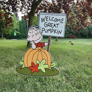 May include: A yard sign featuring a cartoon character with black hair wearing a red and black striped shirt, sitting behind a large orange pumpkin. The character holds a white rectangular sign with the black text WELCOME GREAT PUMPKIN.
