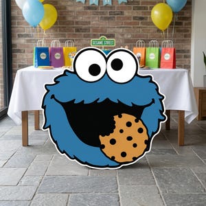 May include: A large, blue Cookie Monster cutout with a cookie in its mouth. The Sesame Street character is in front of a table with colorful gift bags, balloons, and a brick wall backdrop. The sign above the table reads "Sesame Street".