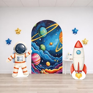 May include: A whimsical space-themed party scene featuring a cardboard astronaut, a rocket ship, and a large arched mural of planets and stars. Metallic star-shaped balloons in gold and blue add to the festive decor. The astronaut is in a white spacesuit with orange accents.