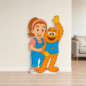 May include: A large cardboard cutout of two cartoon characters. A girl in a pink top and blue dungarees stands beside a furry orange monster wearing a blue top. The characters are smiling and appear friendly.