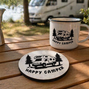 May include: A white enamel mug and wooden coaster with a black camper graphic and the words "Happy Camper." The mug has a black rim and handle. The coaster is round with a black edge. A silver coffee pot sits nearby.