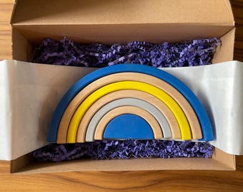 Könnte beinhalten: Ein Holz-Regenbogen-Stapeltier in einem Karton mit lila Füllung. Der Regenbogen hat Schichten aus Blau, Gelb, Naturholz und Grau. Das Spielzeug ist für Kinder konzipiert und ein farbenfrohes Lernwerkzeug.