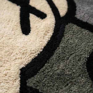 May include: Close-up of a plush rug featuring a graphic design with a repeating pattern. The design incorporates black, off-white, and gray colors. The rug has a soft, textured surface.