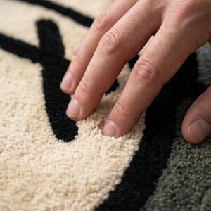 May include: Close-up of a textured rug with a black and off-white design. The rug's surface is soft and plush, with a hand resting on it. The design features bold black lines against a neutral background, creating a modern aesthetic.