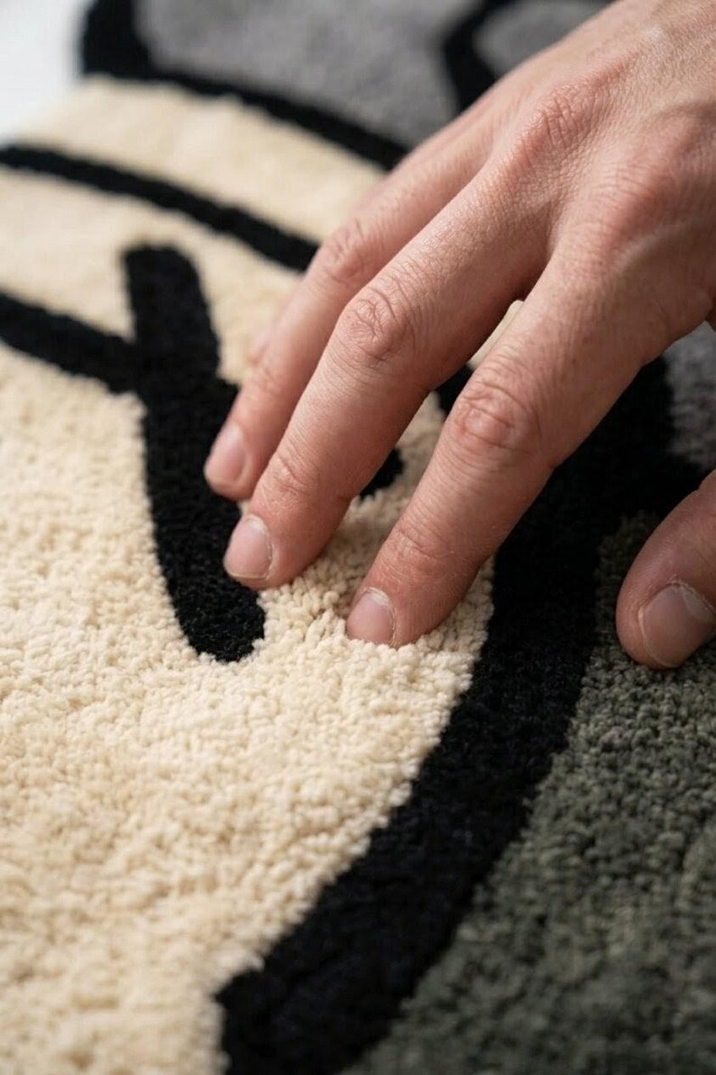 May include: Close-up of a textured rug with a black and off-white design. The rug's surface is soft and plush, with a hand resting on it. The design features bold black lines against a neutral background, creating a modern aesthetic.