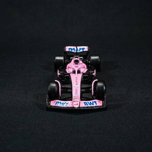 Puede incluir: Un coche de carreras de Fórmula 1 rosa y negro con detalles en azul y blanco. El coche tiene la palabra "Alpine" y "BWT". El coche tiene cuatro ruedas negras. Fondo negro.