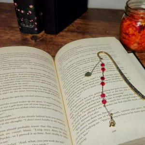 May include: An open book with a decorative bookmark featuring red beads and a small charm. The bookmark is placed on the pages of the book. A jar with orange contents and flower petals are also visible.