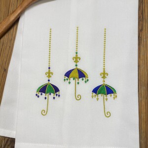 May include: White kitchen towel with embroidered umbrellas in blue, green, and yellow. Each umbrella is suspended by a gold chain and topped with a fleur-de-lis. A wooden spoon and beaded garland are in the background.