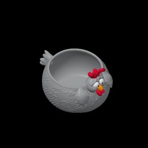 May include: A gray, round, bowl-shaped container designed to resemble a chicken. The exterior features textured, feather-like details, while the front displays a red comb, a yellow beak, a red wattle, and two white eyes with black pupils.