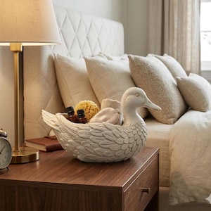 May include: A white ceramic duck-shaped decorative bowl filled with toiletries, including small bottles, a sponge, and a folded towel. The bowl sits on a wooden nightstand next to a lamp and a bed with pillows.