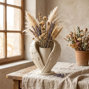 May include: A cream-colored vase shaped like angel wings holds a bouquet of dried flowers, including pampas grass, lavender, and wheat. The vase sits on a wooden table covered with a linen cloth. A terracotta pot with dried flowers is in the background.