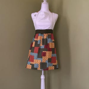 May include: A white apron with a colourful patchwork skirt is displayed on a white mannequin. The skirt features squares of red, blue, yellow, and green fabric. The apron has a dark green waistband and a tag attached to the top.