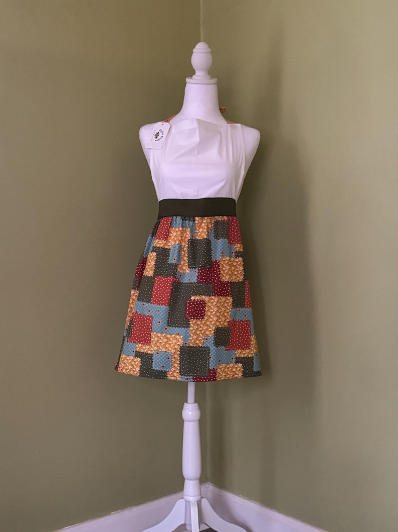 May include: A white apron with a colourful patchwork skirt is displayed on a white mannequin. The skirt features squares of red, blue, yellow, and green fabric. The apron has a dark green waistband and a tag attached to the top.