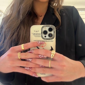 May include: A cream-colored phone case with the text "LA" and a black swoosh design is held up. The person is wearing multiple gold rings and has red and white checkered nail art. A black blazer is in the background.