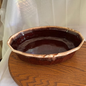 Puede incluir: Una fuente de horno ovalada de cerámica marrón con acabado brillante. La fuente tiene un borde de color claro y está colocada sobre una superficie de madera. El interior de la fuente es de un tono marrón más oscuro.