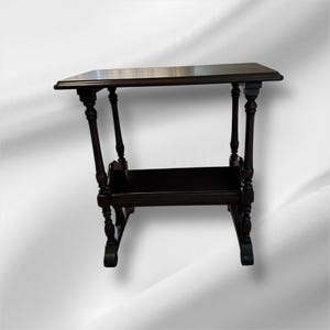 May include: A dark brown, antique side table with a rectangular top and a lower shelf. The table has four ornate, turned legs and a dark finish. The table is approximately 28 inches tall and 24 inches wide.