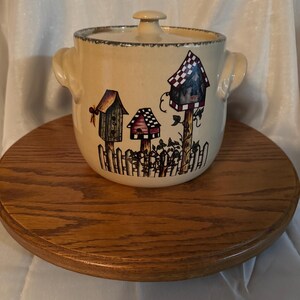 Könnte beinhalten: Beige Keramik-Auflaufform mit Deckel und zwei Griffen. Die Schüssel ist mit einem skurrilen Design von Vogelhäusern, einem weißen Zaun und Blättern verziert. Die Schüssel steht auf einem runden Holzsockel.