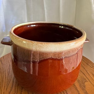 Pode incluir: Uma panela de cerâmica com um esmalte marrom e creme. A panela tem uma abertura larga, duas pequenas alças e uma forma arredondada. O exterior é um marrom rico e brilhante, com uma faixa creme mais clara ao redor da borda.