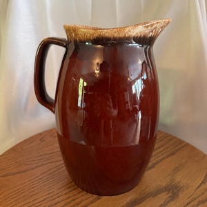 May include: A large, glossy brown ceramic pitcher with a handle. The pitcher has a wide base that tapers to a narrower neck and a flared rim with a lighter brown color. The pitcher is sitting on a wooden surface.