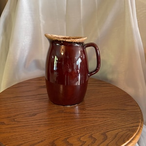 May include: A dark brown ceramic pitcher with a handle. The pitcher has a glossy finish and a lighter brown rim. It is sitting on a round wooden table.