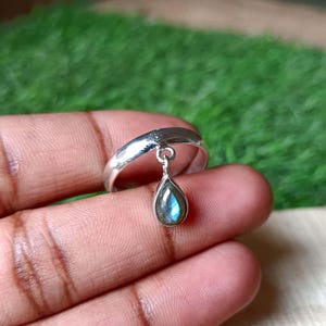 Puede incluir: Un anillo de plata con un colgante de piedra preciosa de labradorita en forma de lágrima. La banda del anillo es lisa y pulida, y la piedra preciosa muestra tonos iridiscentes azules y verdes. El anillo se sostiene en una mano sobre un fondo verde borroso.
