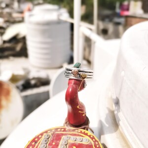 Puede incluir: Un anillo de plata con tres bandas, con dos pequeños acentos de corazón en oro. El anillo se muestra sobre un objeto decorativo rojo con patrones amarillos y rojos, sobre un fondo borroso al aire libre.