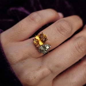 Könnte beinhalten: Ein Goldring mit zwei verschiedenen Steinen, die nebeneinander angeordnet sind. Ein Stein ist ein facettierter, oval geschliffener gelber Citrin, der andere ein strukturierter, metallisch grauer Pyritstein. Der Ring ist dünn und golden.