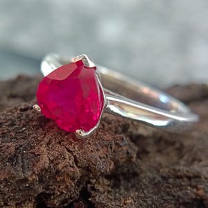 May include: A silver ring featuring a vibrant, heart-shaped, red gemstone. The gemstone is the focal point, set in a simple, elegant band. The ring is displayed on a textured, brown surface, highlighting the contrast between the metal and the stone.
