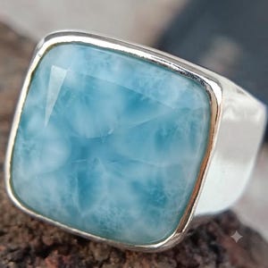 May include: A square-shaped ring featuring a polished silver band and a large, smooth, light blue Larimar gemstone with white cloud-like patterns. The stone is set in a simple silver bezel, highlighting the natural texture and color of the stone.