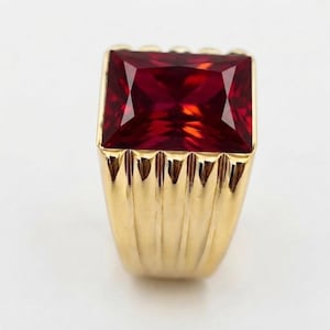 May include: A gold ring featuring a large, rectangular, deep red gemstone set in a prominent, faceted cut. The band of the ring is crafted from polished yellow gold and displays a series of vertical, grooved channels along its outer surface.