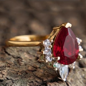 Può includere: Un anello in oro con una grande pietra preziosa rossa a forma di pera come elemento centrale, circondata da un gruppo di piccole pietre d'accento chiare a taglio rotondo. La fascia è in oro liscio e lucido, in contrasto con la pietra.