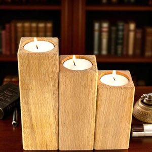 May include: Three wooden candle holders of varying heights, each with a lit white candle. The holders are rectangular blocks of light-colored wood. The background includes a bookshelf and desk accessories.