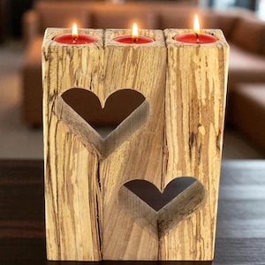 May include: A wooden candle holder with three red candles. The holder is made of light-colored wood with heart-shaped cutouts. The candles are lit, casting a warm glow. The wood has a natural, textured appearance.
